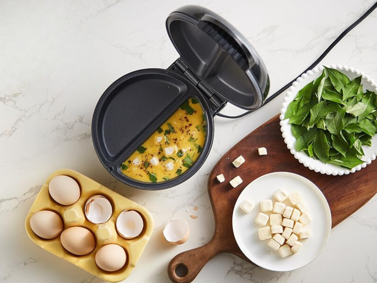 An omeltte maker on a kitchen counter with eggs and ingredients.